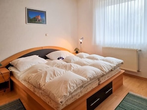 Ein Schlafzimmer mit Bett, Nachttisch, Lampe, Fenster mit Vorhängen, Heizkörper und einem gerahmten Bild an der Wand.