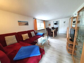 Ein Wohnzimmer mit rotem Sofa, einem Glastisch, einem Essbereich mit Holztisch und Stühlen sowie einem Fernseher auf einem Holzschrank.