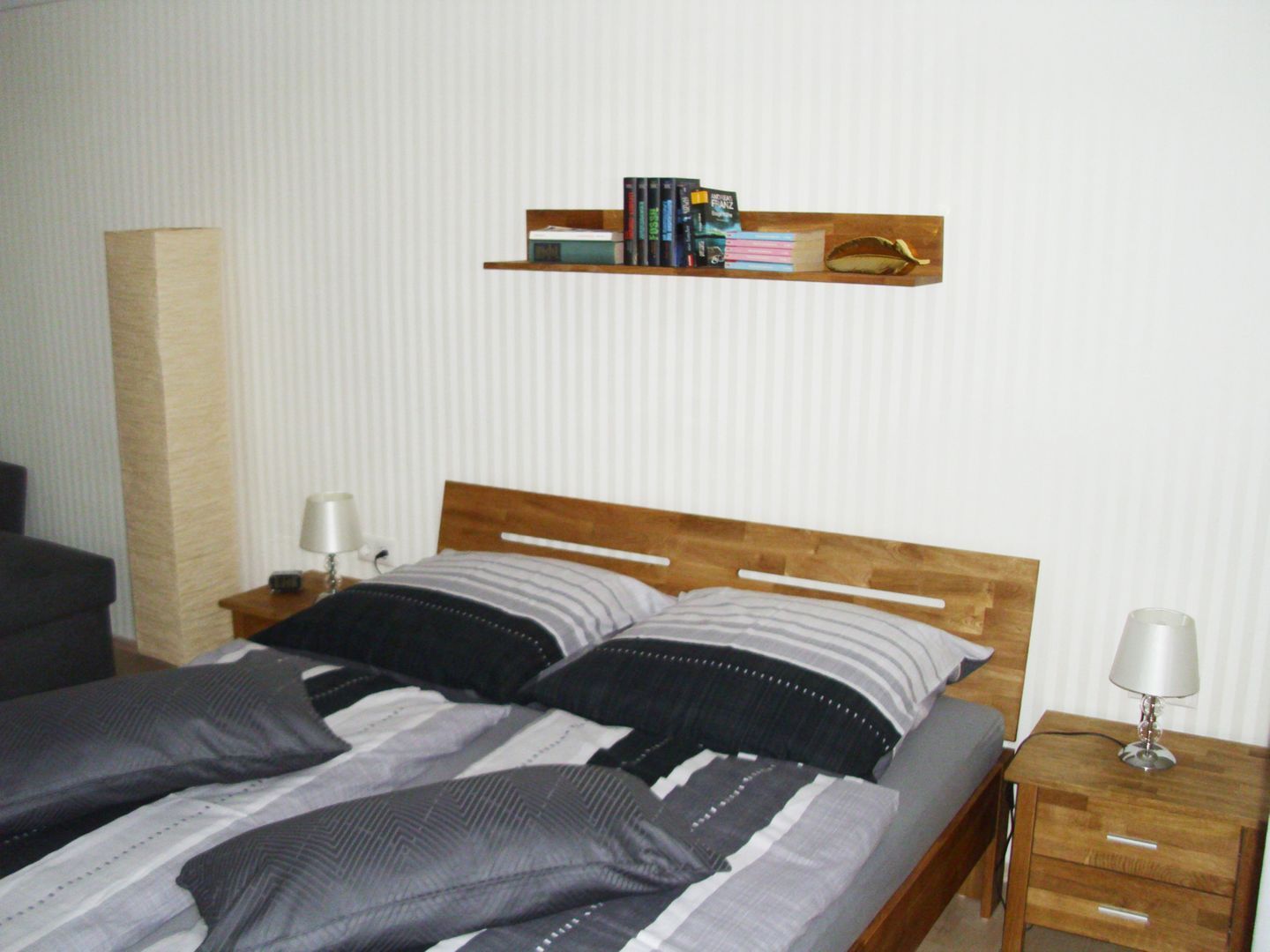 Een tweepersoonsbed met gestreept beddengoed, een houten hoofdbord, nachtkastjes met lampen, een boekenkast vol boeken en een hoge, cilindervormige object aan de muur.
