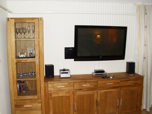A living room with a flat-screen TV mounted on the wall, wooden cabinets below, and a shelf with decorative items. - Vacation apartment on the outskirts (Sassnitz auf Rügen)