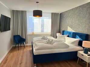 A modern hotel room with a bed, a chair, a bedside table with a lamp, and a window with curtains. - Vacation apartment Little Sea Lion (Sassnitz)