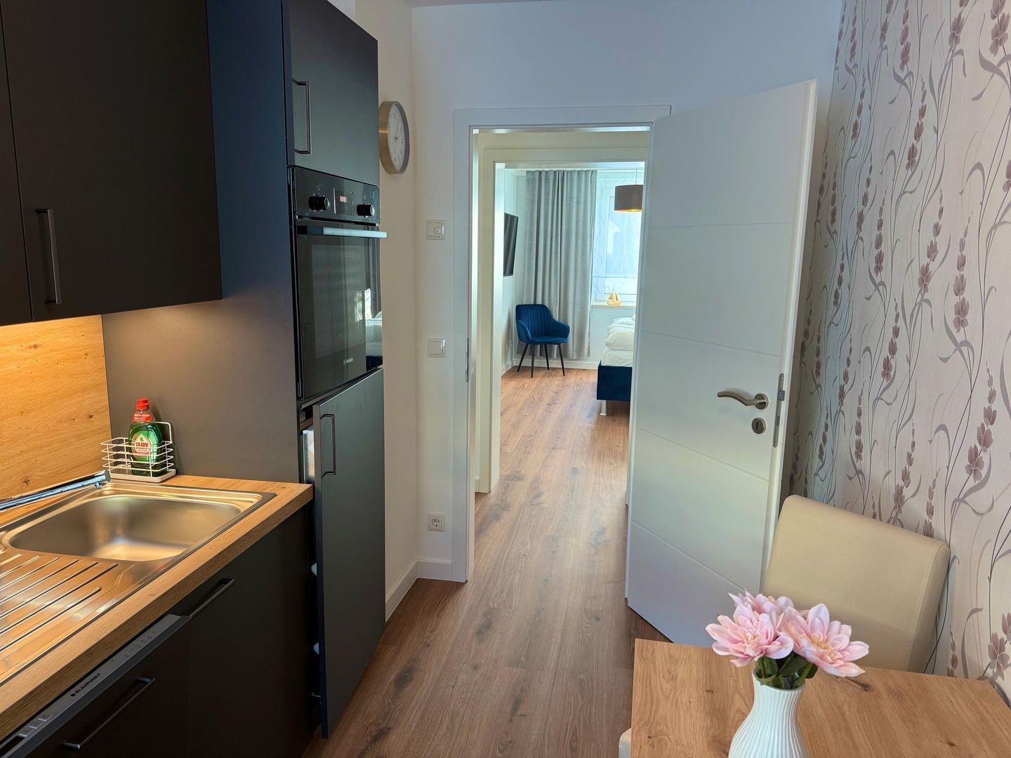 Una cocina moderna con horno empotrado y fregadero, un comedor con mesa de madera y un jarrón de flores, y un dormitorio visible a través de una puerta abierta.