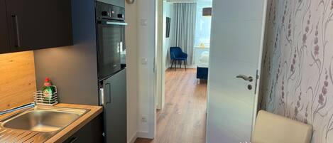 Una cocina moderna con horno empotrado y fregadero, un comedor con mesa de madera y un jarrón de flores, y un dormitorio visible a través de una puerta abierta.