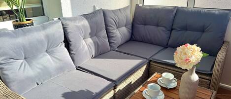 A wicker sofa with grey cushions, a small wooden table with cups and a vase, and a potted plant.