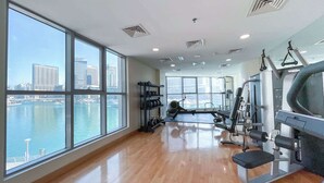 Ett gym med stora fönster som vetter mot en sjö eller ett hav och en stadsbild, trägolv och diverse träningsredskap.