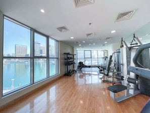 Ein Fitnessstudio mit großen Fenstern, die auf ein Gewässer und eine Stadtansicht blicken, Holzböden und unterschiedlicher Trainingsausstattung.