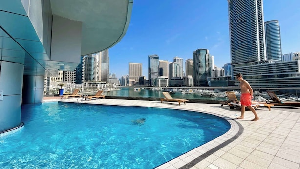 En pool med solstolar, en stadsbild i bakgrunden och en modern byggnad.