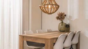 Zona pranzo con tavolo in legno, sedie bianche, lampadario e un vaso con fiori secchi.