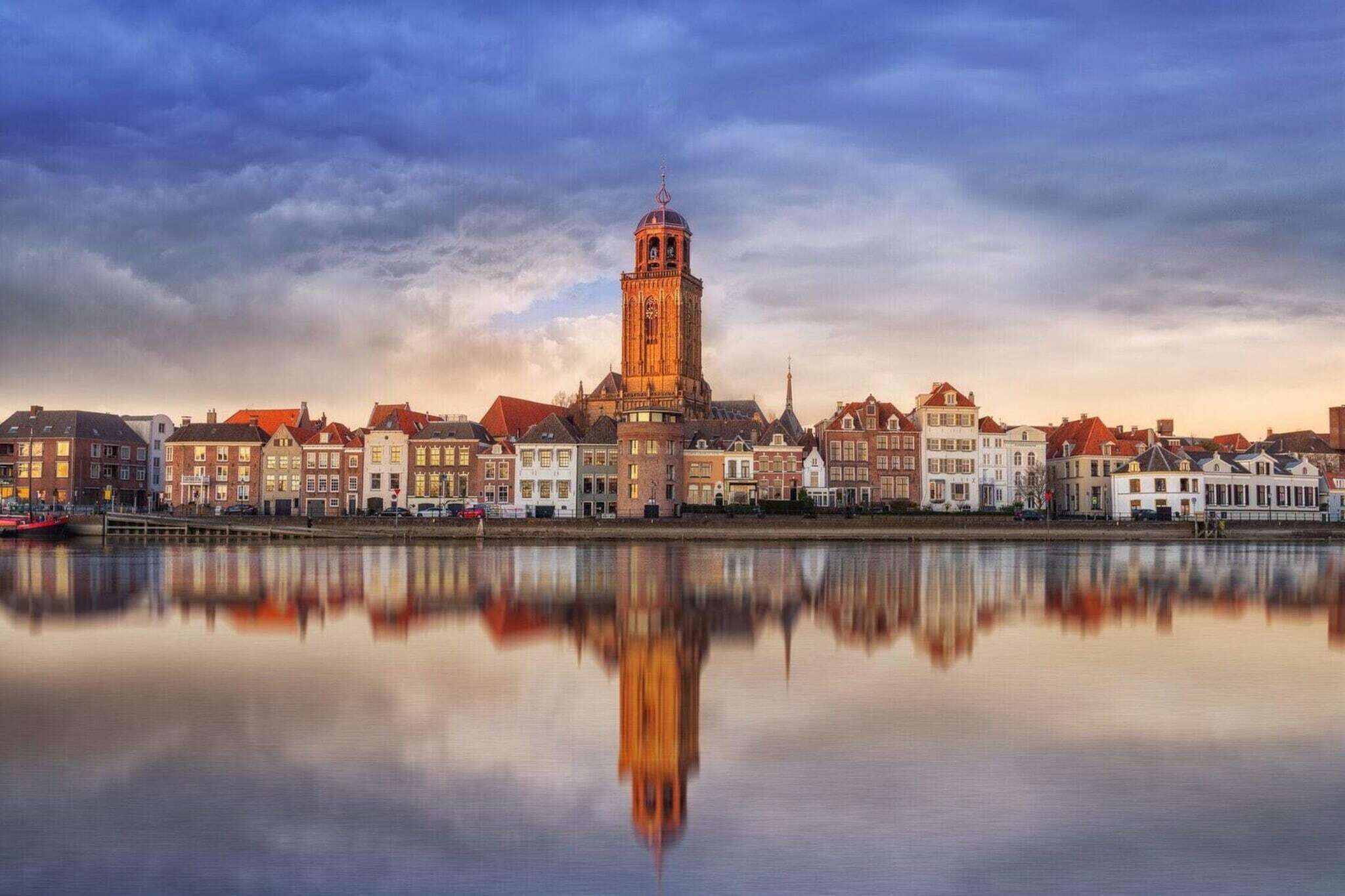 En historisk by med et markant tårn, der spejler sig i rolige vand, omgivet af traditionelle bygninger.