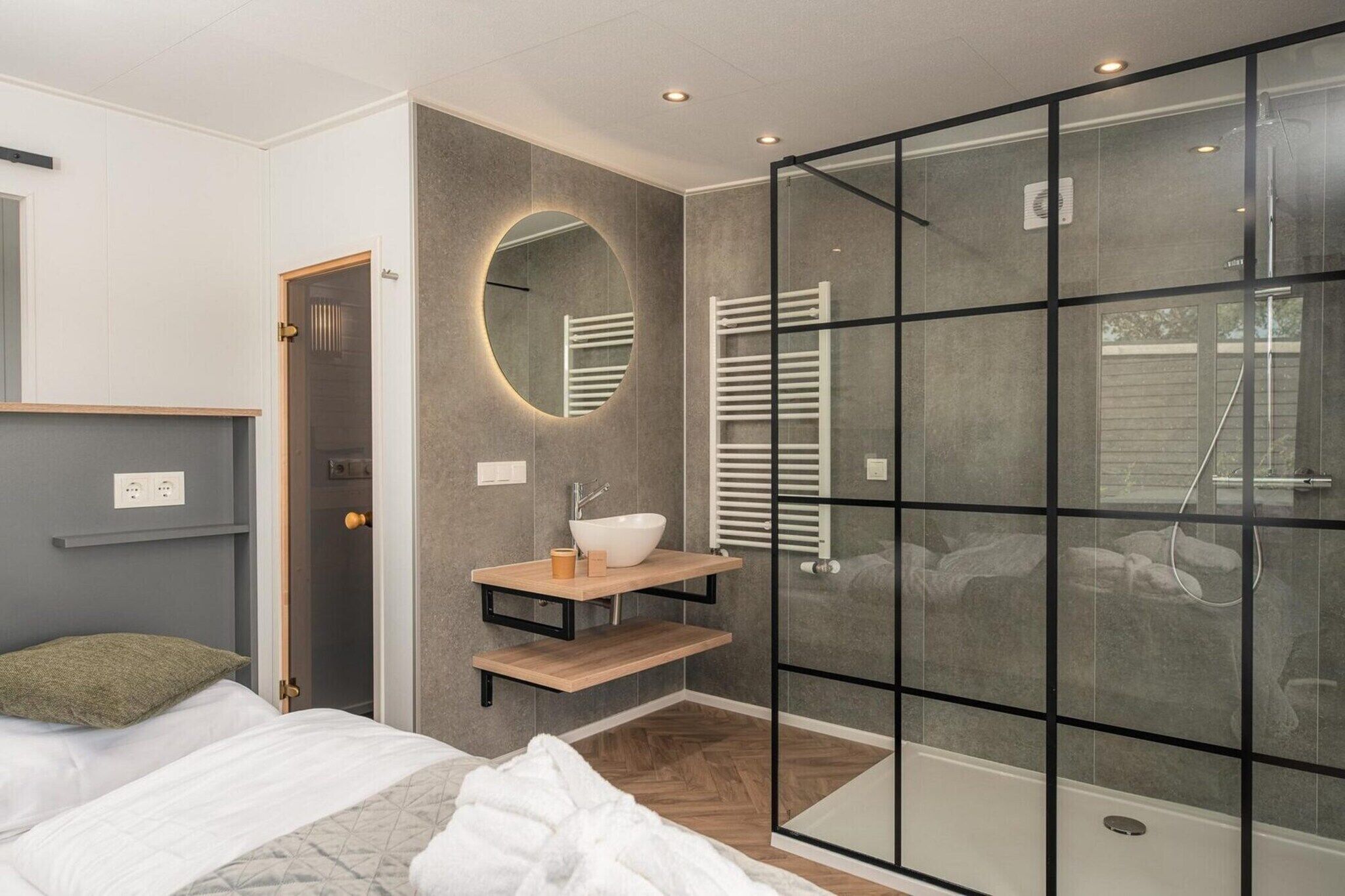 A modern bedroom with a bed, a bedside table, a mirror, a built-in shelf, a glass shower enclosure, and a door leading to another room.