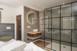Een moderne slaapkamer met een bed, nachtkastje, spiegel, inbouwkast, douchecabine van glas en een deur naar een andere kamer.