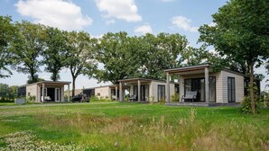 Uma fileira de cabanas modernas, térreas, com varandas abertas, cercada por um campo gramado e árvores.