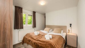 A bedroom with a bed, a window with curtains, a bedside table with a lamp, and a radiator. - Pet-Friendly Forest Lodge with Fenced Garden (Rijssen)