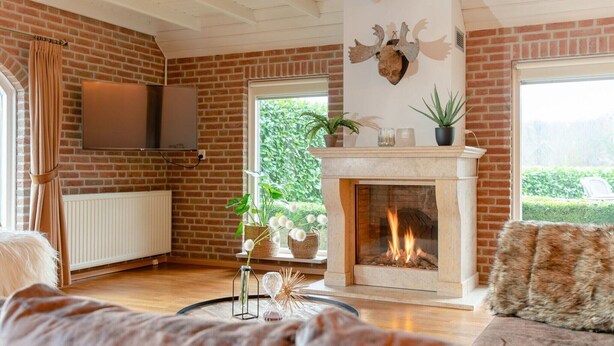 A living room with a fireplace, brick walls, a mounted TV, and a view of greenery outside. - Retreat with Authentic Thatched Roof (Zuna)
