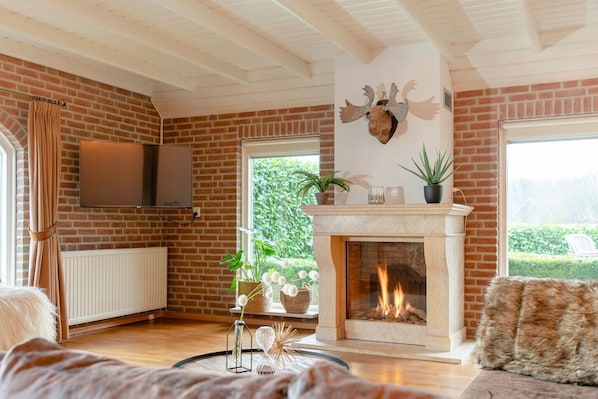 Una sala de estar con chimenea, paredes de ladrillo, un televisor montado y vista a espacios verdes exteriores.
