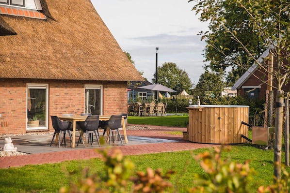 A brick building with a thatched roof, a wooden hot tub, and a table with chairs in the foreground.
