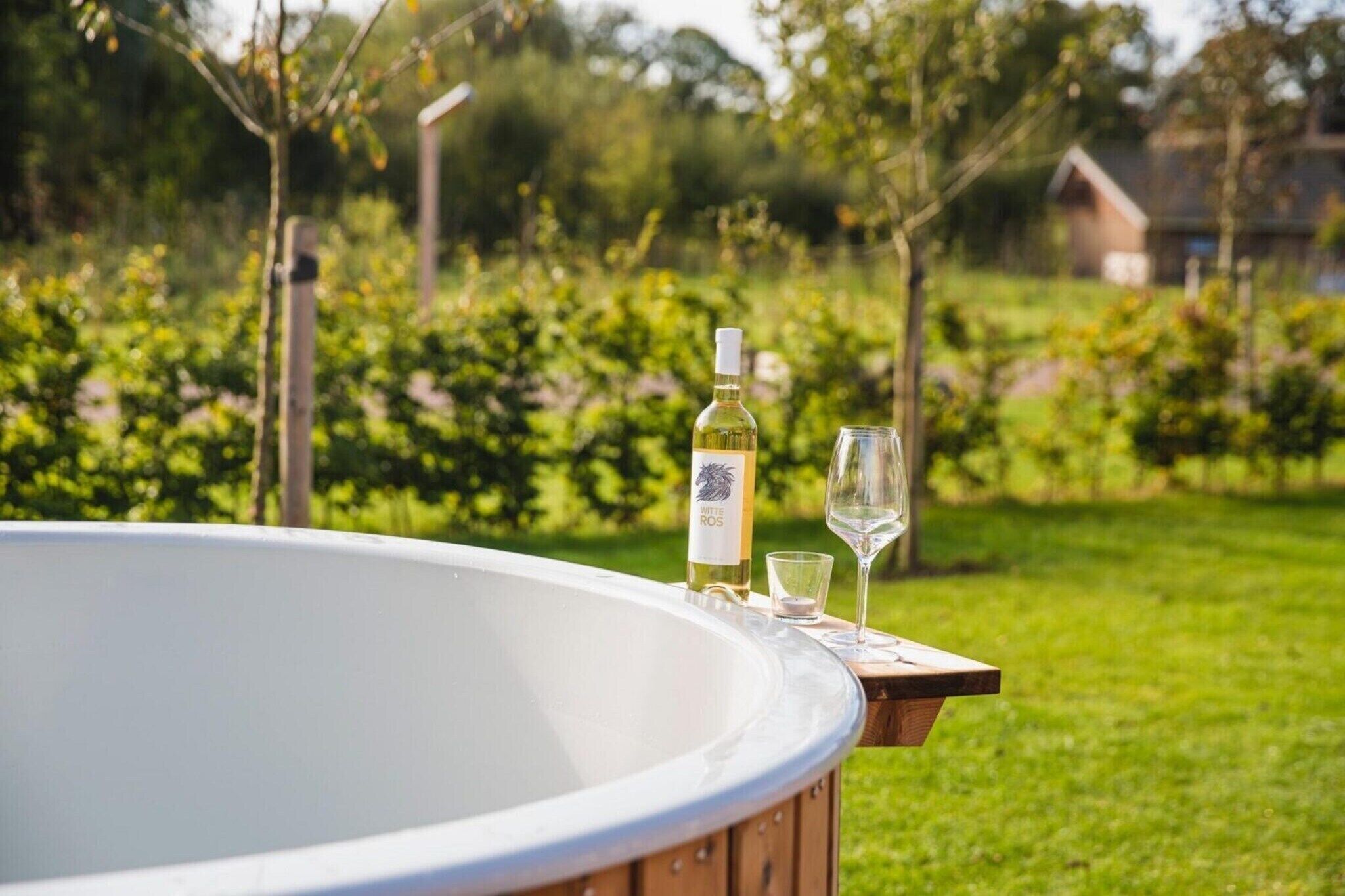 A hot tub with a bottle of white wine and a glass on a wooden platform, set against a vineyard backdrop.