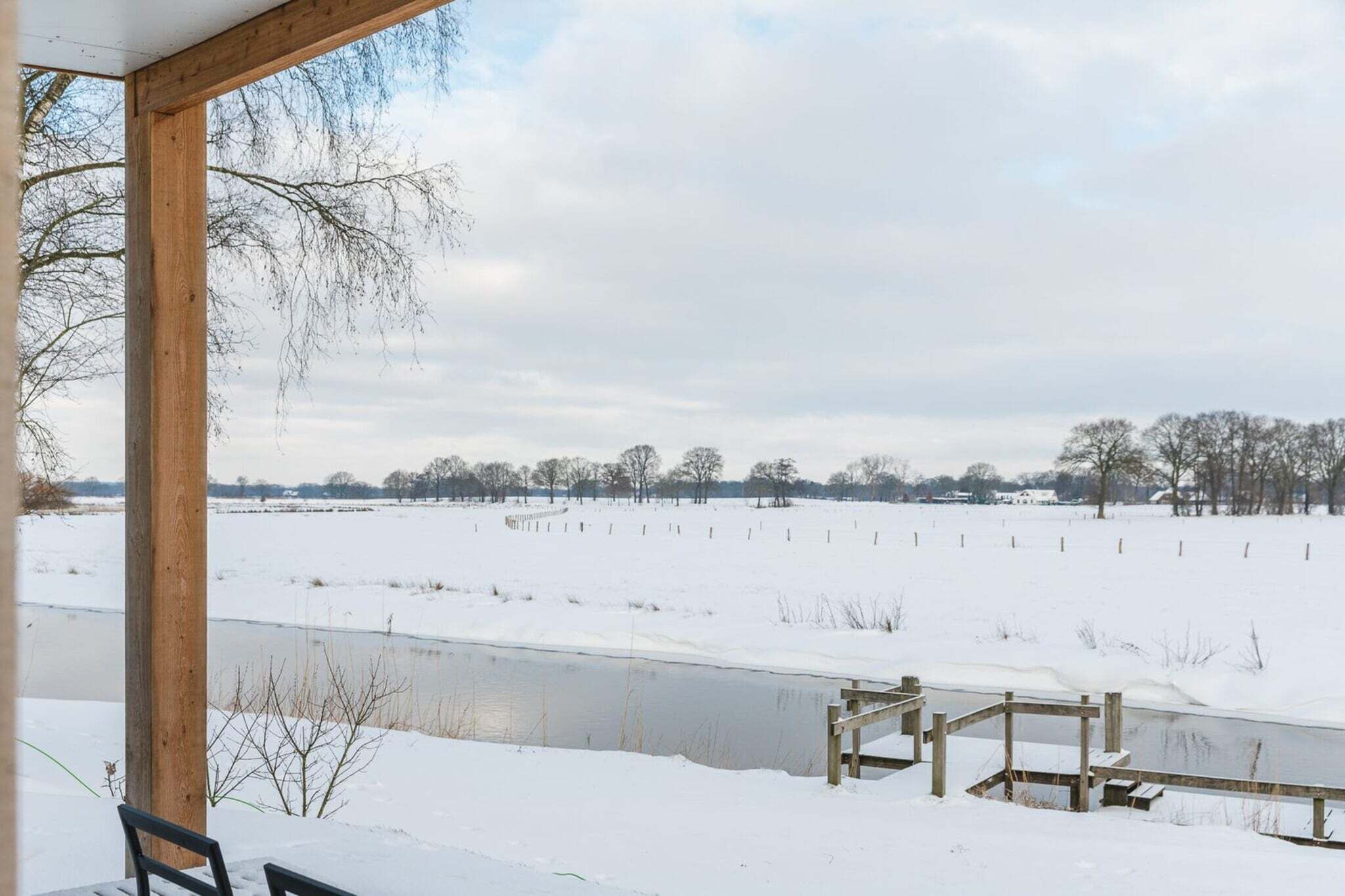 Uitzicht vanuit een overdekt buitengebied met een houten constructie, een met sneeuw bedekte weide en een waterlichaam.