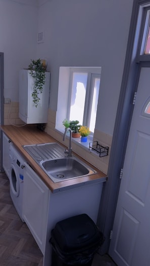 Een kleine keuken met een spoelbak, een wasmachine, een raam met planten en een deur.