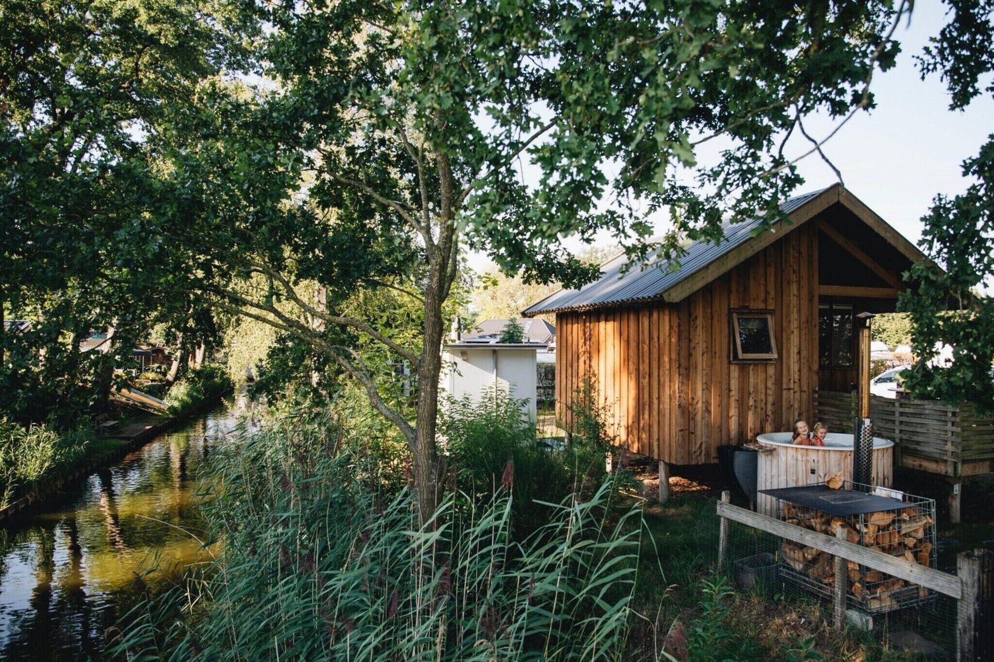 A rustic wooden cabin with a hot tub, surrounded by lush greenery and a calm river.