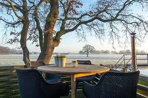 A table with chairs, a potted plant, and a view of a body of water with trees and a misty landscape.