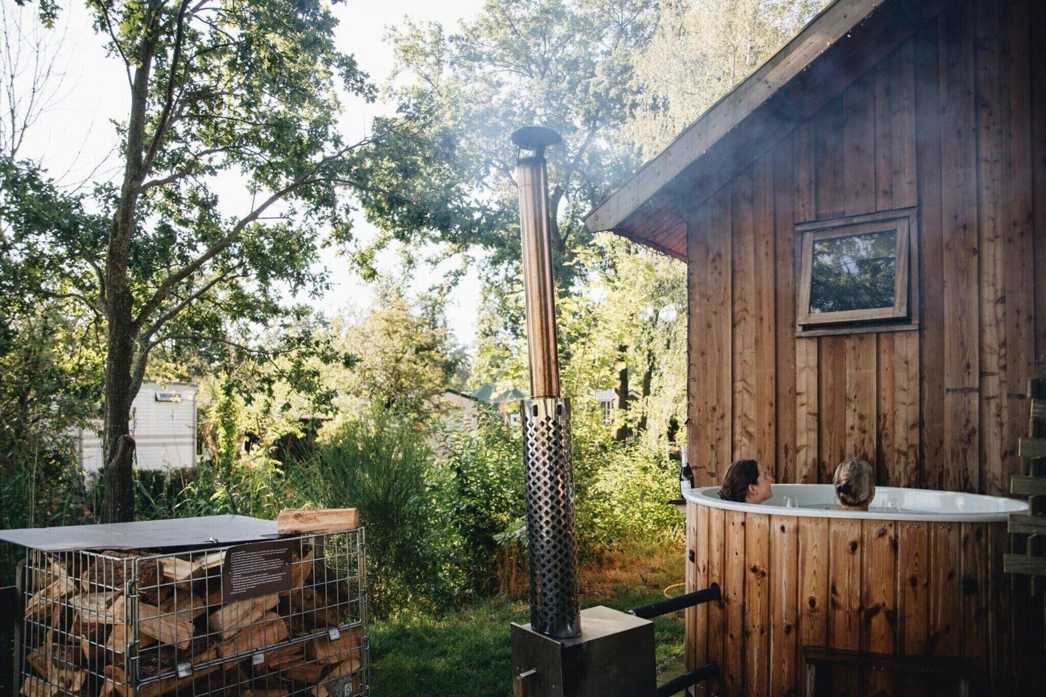 Una cabaña de madera con hidromasaje, una parrilla metálica y leña apilada.