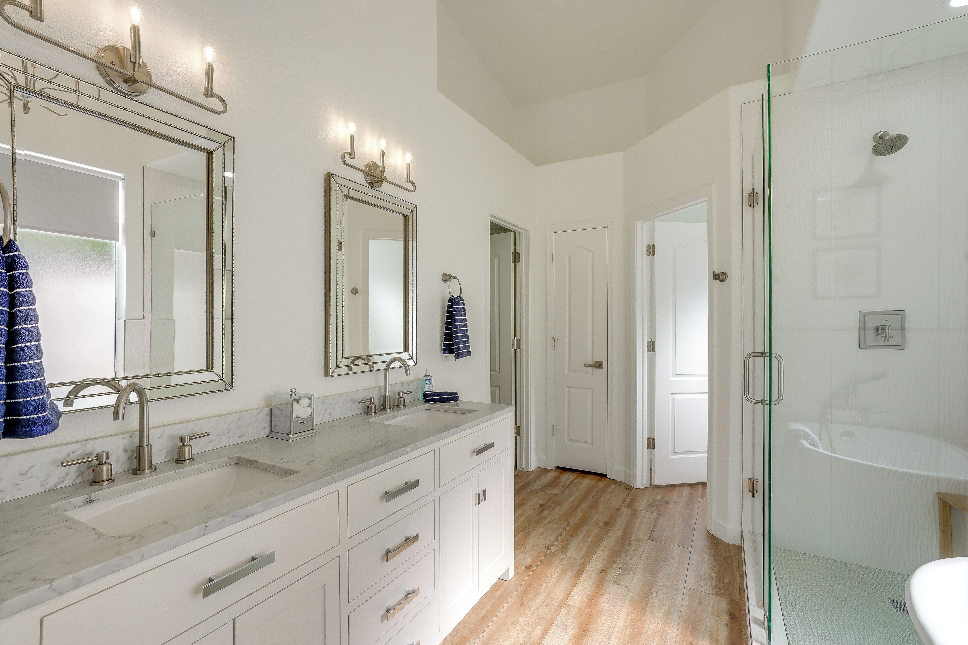 A bathroom with a large mirror, two sinks, a shower, and a door leading to another room.
