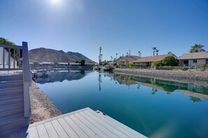 A tranquil water body with a wooden deck, reflecting nearby buildings and palm trees, under a clear sky.