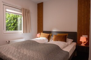 Een keurig opgemaakt bed met een gewatteerde spreidek, een raam met gordijnen, een bedlampje en een houten hoofdbord.