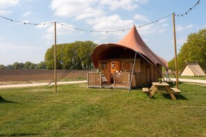 A wooden tent with a conical roof, a porch with a bench, and a picnic table. - Glamping Oasis with Bohemian Flair (Zuna)