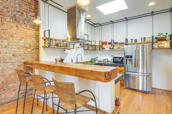 Una cocina con barra de madera, sillas de mimbre, un refrigerador de acero inoxidable y una pared de ladrillo.