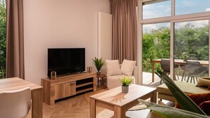 A modern living room with a flat-screen TV, wooden furniture, a sofa, and a large window with a view of greenery.