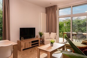 Ett modernt vardagsrum med en platt-TV, trämöbler, en soffa och ett stort fönster med utsikt över grönområden.