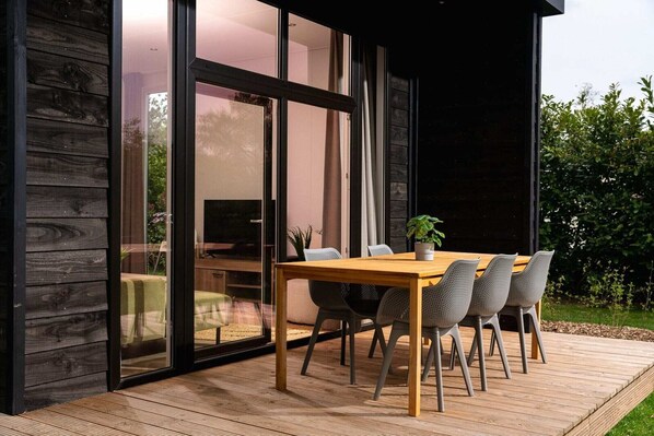 A modern house with a wooden deck, a dining table with chairs, and a large glass door leading to an interior space.