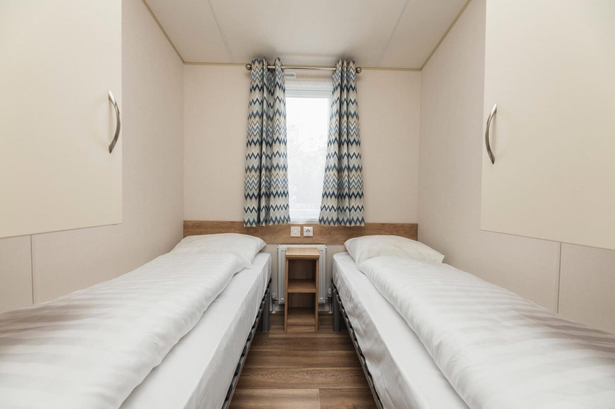 Een kamer met twee éénpersoonsbedden, een klein tafeltje en een raam met gordijnen met een patroon.