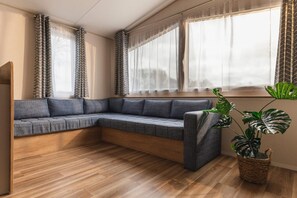 Um sofá de canto com base de madeira, uma planta em vaso e uma janela com cortinas leves.