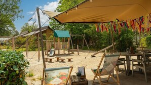 Un espacio al aire libre rústico con mesas y sillas de madera, una zona de sombra con una carpa y una pequeña estructura de juegos.