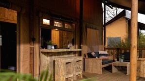 Una terraza de madera con sofá y mesa, una zona de bar con taburetes y un ventanal con vistas a la vegetación.