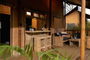 Eine Holzterrasse mit einer Couch und einem Tisch, eine Bar mit Hockern und ein Fenster mit Blick auf Grünflächen.