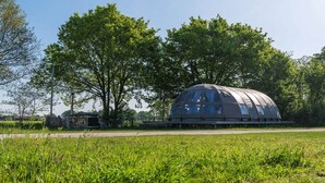 A dome-shaped structure with a transparent roof, surrounded by a grassy field and trees. - Unique Riverdome Stay with Hot Tub (Zuna)