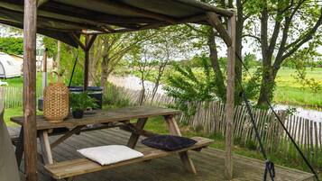 A wooden picnic table with cushions, a woven basket, and a black object on a deck overlooking a river.