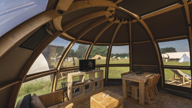 A dome-shaped interior with a wooden table, chairs, a TV, and large windows offering a view of tents outside. - Unique Riverdome Stay with Hot Tub (Zuna)