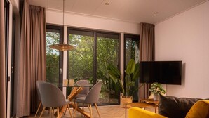 A modern living room with a sofa, TV, dining table, chairs, and large windows. - Home near National Park (Zuna)