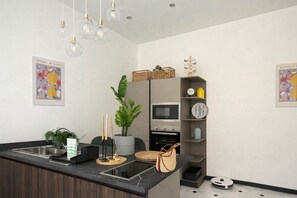 Eine moderne Küche mit einer zentralen Kochinsel, einer Mikrowelle, einem Spülbecken und einem kleinen Backofen. Deko-Elemente wie eine Pflanze, Kerzen und ein Korb sind vorhanden.