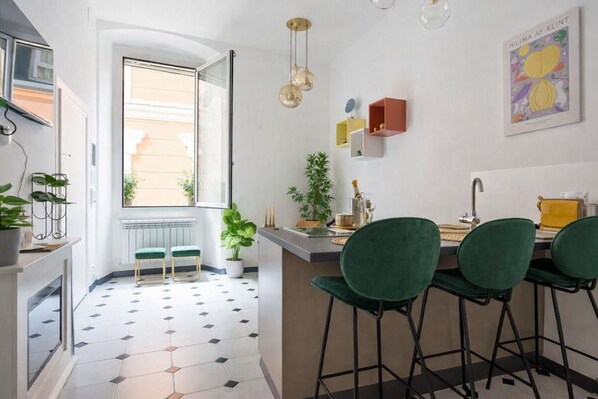 A modern kitchen with a dining area, featuring a large window, green chairs, and a minimalist design.