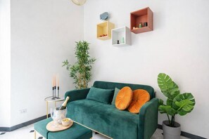 A green sofa with orange cushions, a small table with a bottle and glasses, and a potted plant.
