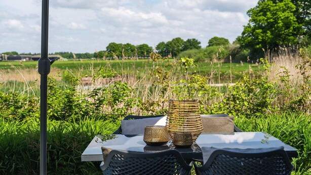 Een buitentafel met een geweven vaas en kaars, onder een luifel, met uitzicht op een grasveld.