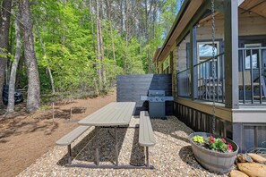 Une terrasse en bois avec une table de pique-nique et un barbecue, entourée d’arbres.