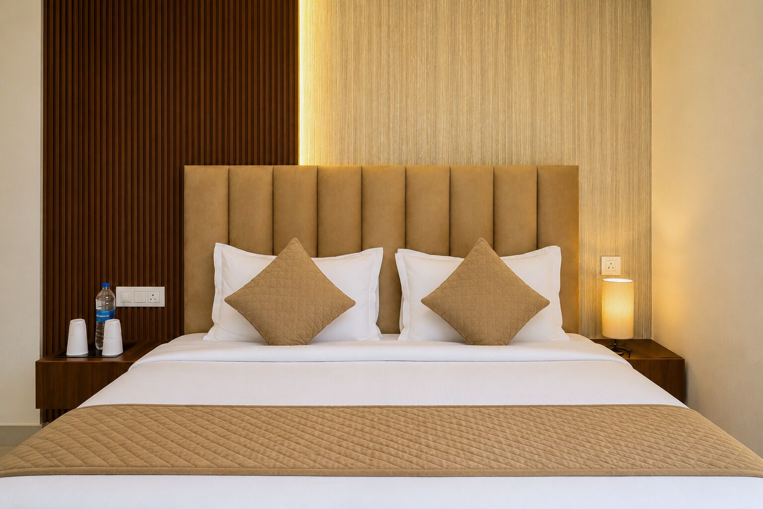 Habitación de hotel con una cama grande, dos almohadas, una mesita de noche con lámpara y una botella de agua.