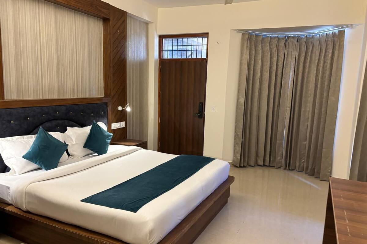 Una habitación de hotel con una cama grande, un cabecero de madera, una mesita de noche, un escritorio de madera, una ventana con cortinas y una puerta con ventanal.
